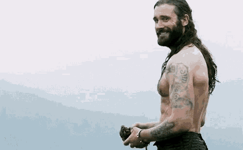 a shirtless man with long hair and tattoos is standing in a field holding a sword .