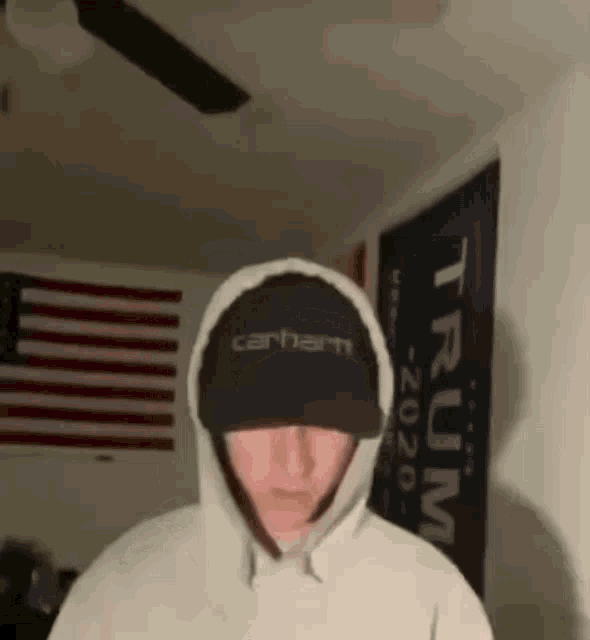 a man wearing a hoodie and a carhartt hat stands in front of a trump sign