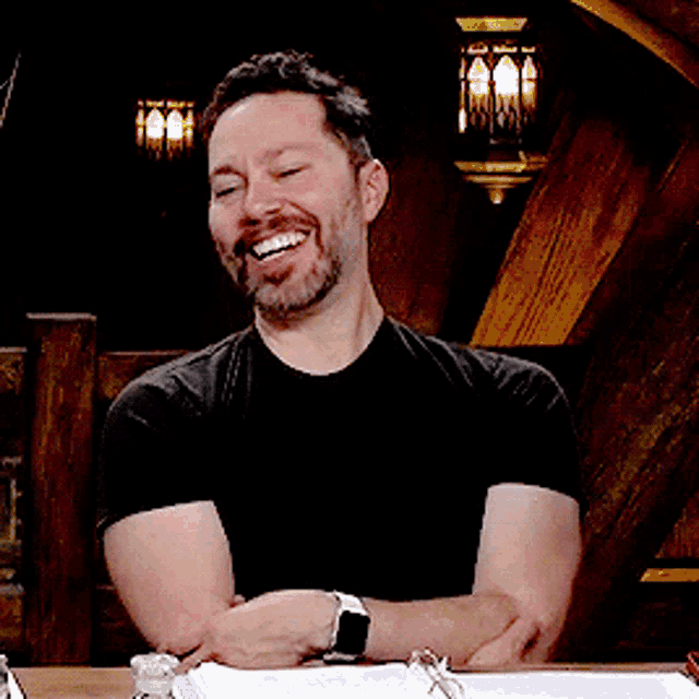 a man in a black shirt is sitting at a table with his arms crossed and a watch on his wrist