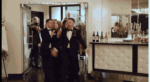 a group of men in tuxedos are dancing in a room with bottles of wine