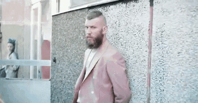 a man with a beard wearing a pink suit is leaning against a concrete wall .