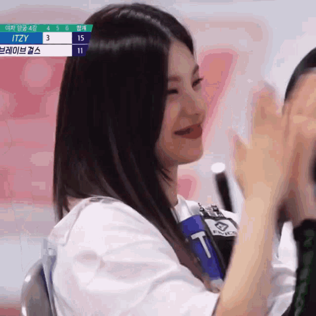 a girl with long black hair is clapping her hands in front of a scoreboard that says itzy