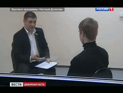 a man in a suit is talking to another man in front of a tv screen that says " вести "