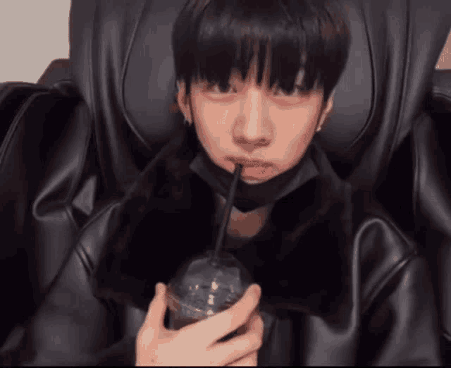 a young man wearing a black mask is sitting in a chair drinking from a plastic cup with a straw .
