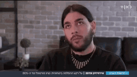 a man with a beard and earrings is on a television screen with a brick wall in the background