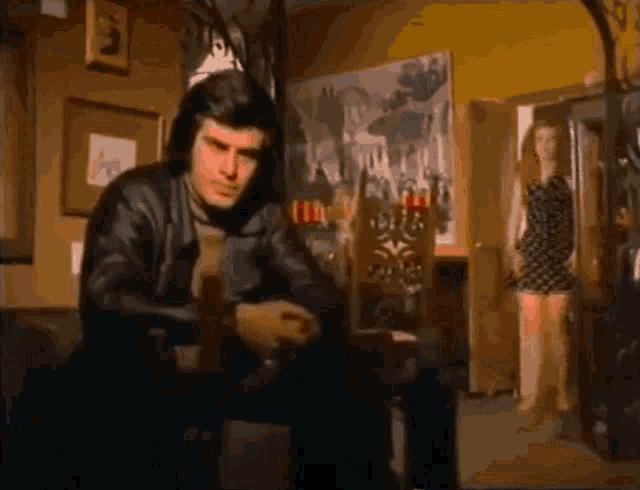 a man in a leather jacket sits in a chair in a room with a woman standing in the background