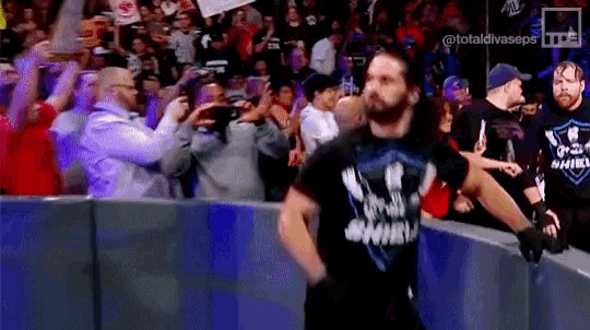 a man in a shield shirt is walking through a wrestling ring while a crowd watches .