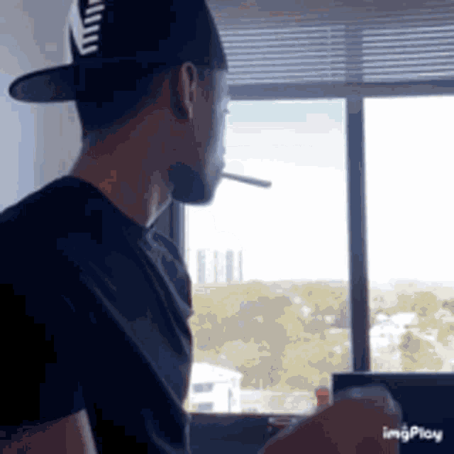 a man wearing a hat is smoking a cigarette in front of a window .