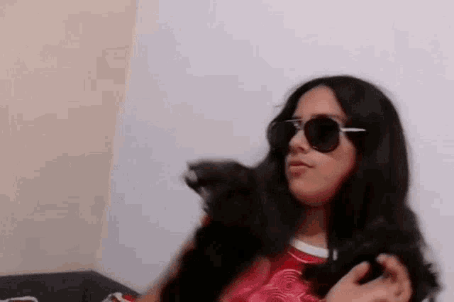 a woman wearing sunglasses is sitting on a couch holding a black cat .