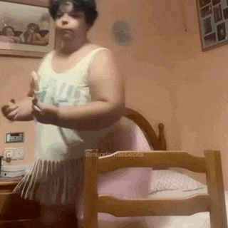 a woman in a white tank top and pink skirt is standing in a bedroom next to a chair .
