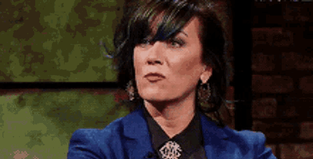 a woman in a blue suit and black shirt is sitting in front of a brick wall and making a funny face .