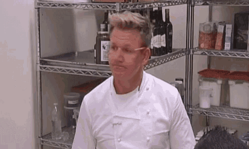 a man in a white chef 's jacket is standing in front of a shelf filled with bottles of wine .