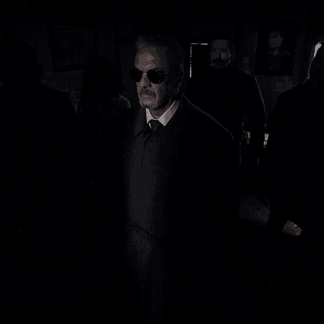 a man wearing sunglasses and a brown coat is standing in a dark room