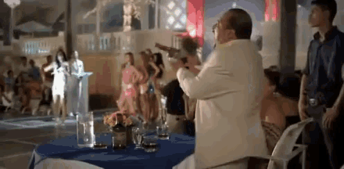 a man in a white suit is holding a gun in front of a group of people at a party .