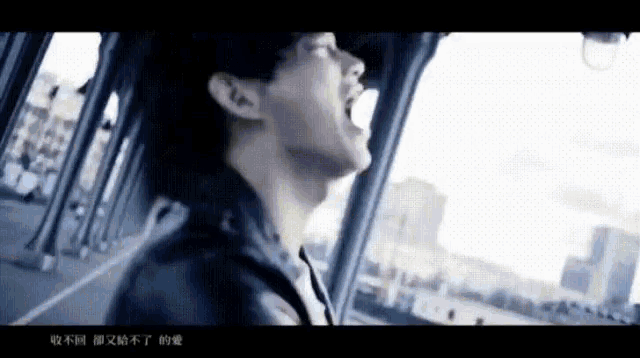 a man in a black jacket is singing into a microphone while looking out a window