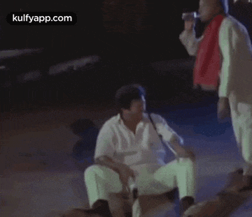 a man is sitting on the ground with a gun in his hand while another man walks by .