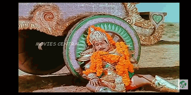 a man in a costume with flowers around his neck is laying on the ground in front of a vehicle .