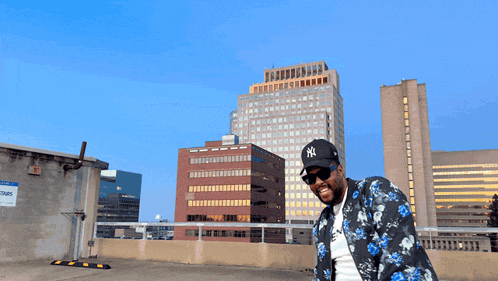 a man wearing a ny hat and sunglasses stands in front of a large building