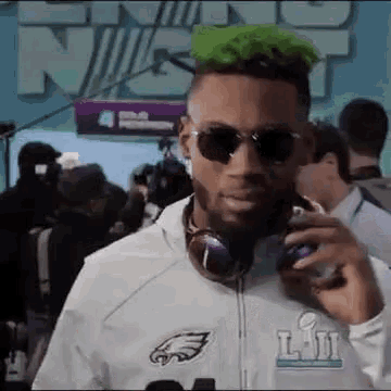 a man with green hair is talking on a cell phone while wearing sunglasses and headphones .