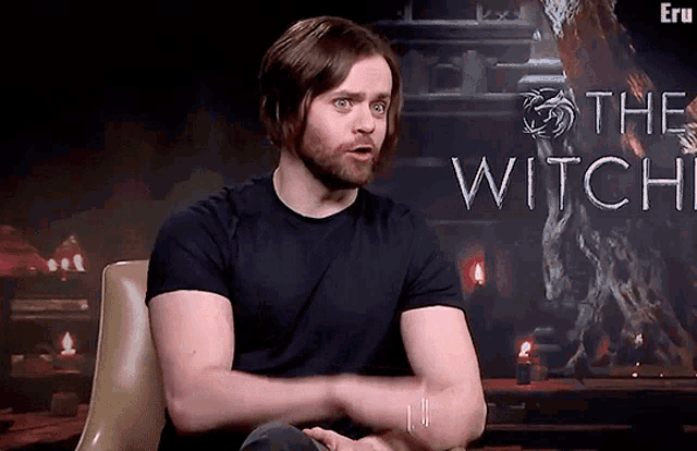 a man in a black shirt is sitting in front of a sign that says " the witch "