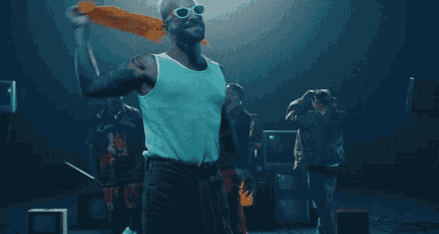 a man wearing sunglasses and a white tank top stands in front of a group of people