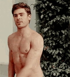 a shirtless man with a tattoo on his arm stands in front of a plant