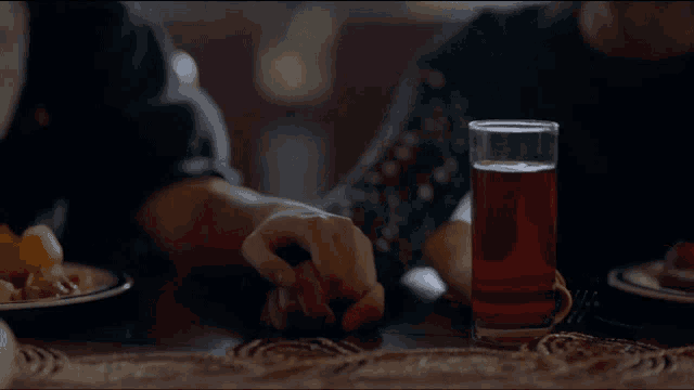 a glass of red liquid sits on a table next to a person 's hand