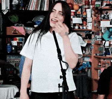 a woman in a white shirt stands in front of a microphone in a room filled with lots of toys and books