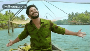 a man in a green shirt is sitting on a boat in the water with his arms outstretched .