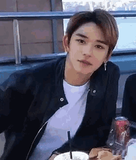 a young man in a black jacket is sitting at a table with a drink and a can of soda .