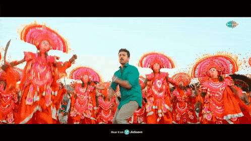 a man in a green shirt is dancing in front of a crowd of people dressed in orange and pink costumes .