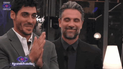 two men are clapping in front of a sign that says grande fratello on it
