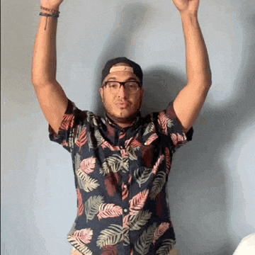 a man wearing a hawaiian shirt and a baseball cap is holding his arms in the air .