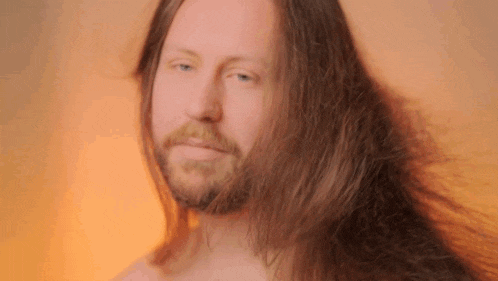 a shirtless man with a beard and long hair looks at the camera