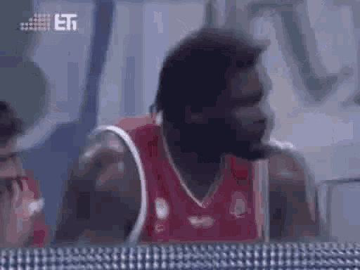a man in a red basketball jersey is talking on a cell phone while sitting on a bench .