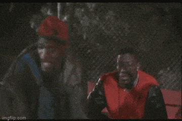 two men are sitting on a bench in front of a chain link fence . one is wearing a red jacket .