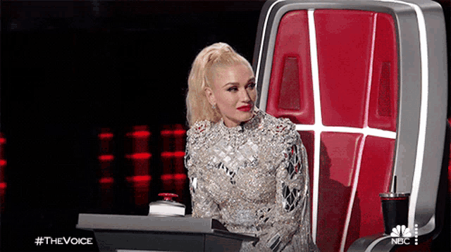 a woman in a silver dress is sitting in a chair with a nbc logo in the background