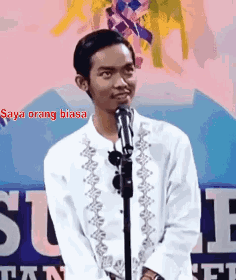 a young man stands in front of a microphone with the words saya orang biasa behind him