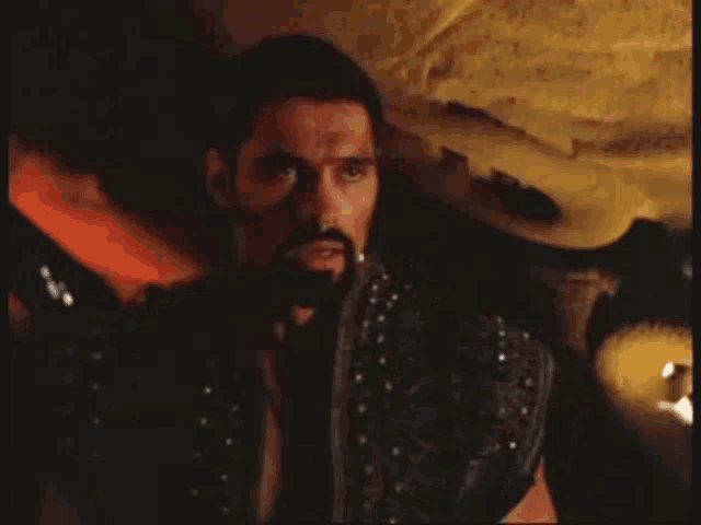 a man with a beard and studded jacket is standing in a dark room