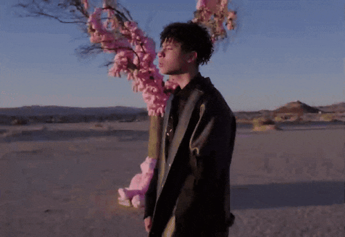a man in a black coat stands in the desert holding a pink teddy bear