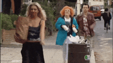 a group of people are walking down a street with a trash can that says ' recycle ' on it .