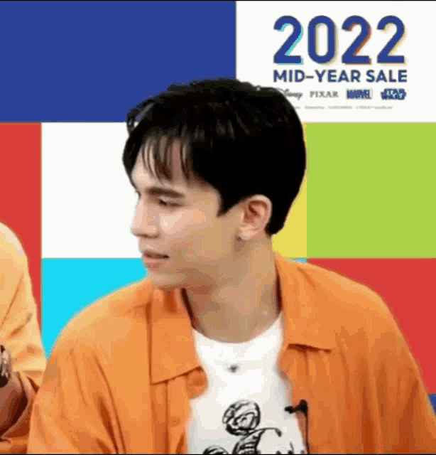 a man in an orange shirt is sitting in front of a sign that says `` 2022 mid-year sale '' .