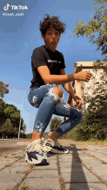 a young man wearing ripped jeans and a black t-shirt is squatting down on a brick sidewalk .