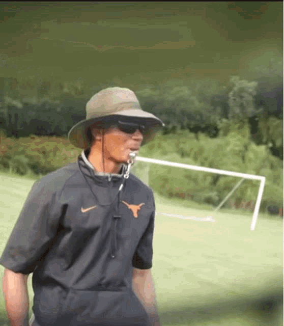 a man wearing a hat and sunglasses is standing in a field with a soccer goal in the background