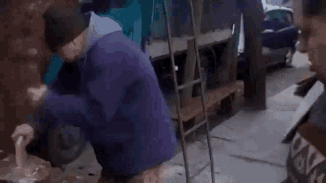 a man in a purple jacket is kneeling down next to a tree stump with a hammer .