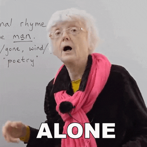 an older woman wearing glasses and a pink scarf says alone in front of a white board