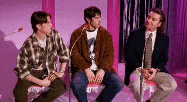 three men are sitting in front of a microphone in a pink room