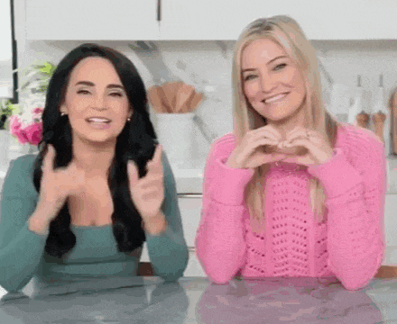 two women are sitting at a table and making a heart shape with their hands .