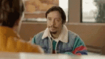 a man in a colorful jacket is sitting at a table talking to another man in a diner .