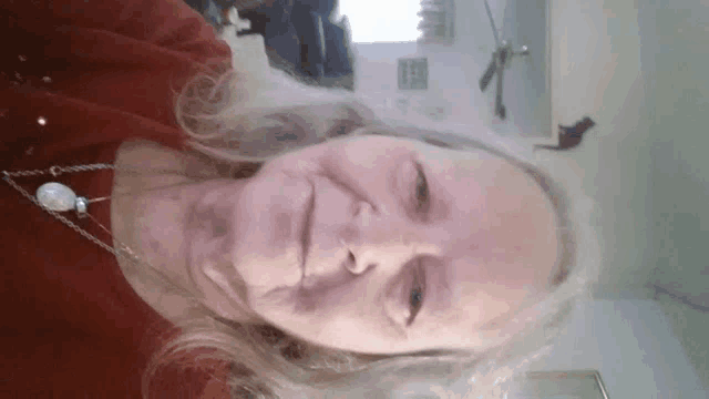 an older woman wearing a red sweater and a pearl necklace looks at the camera